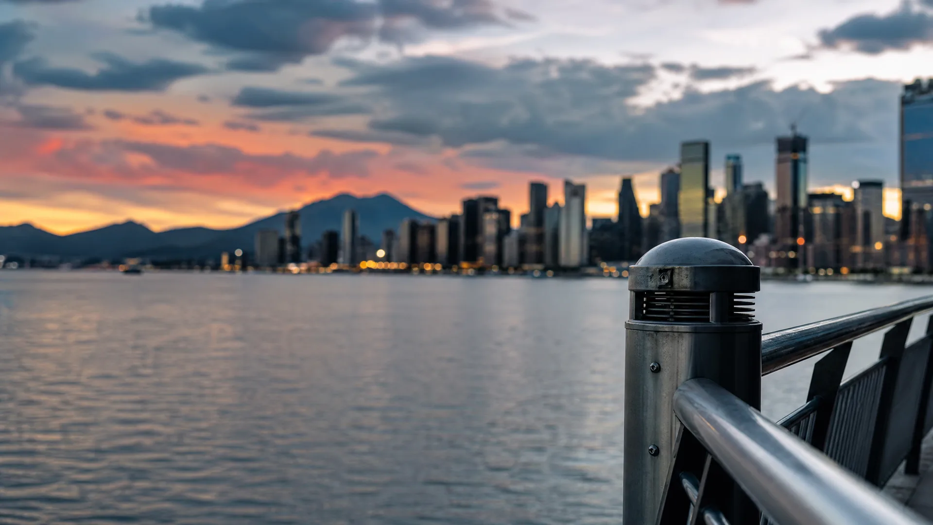 A metal railing and domed bollard light in sharp focus along a waterfront, with a blurred city skyline, distant mountains and orange sunset clouds across the water.
